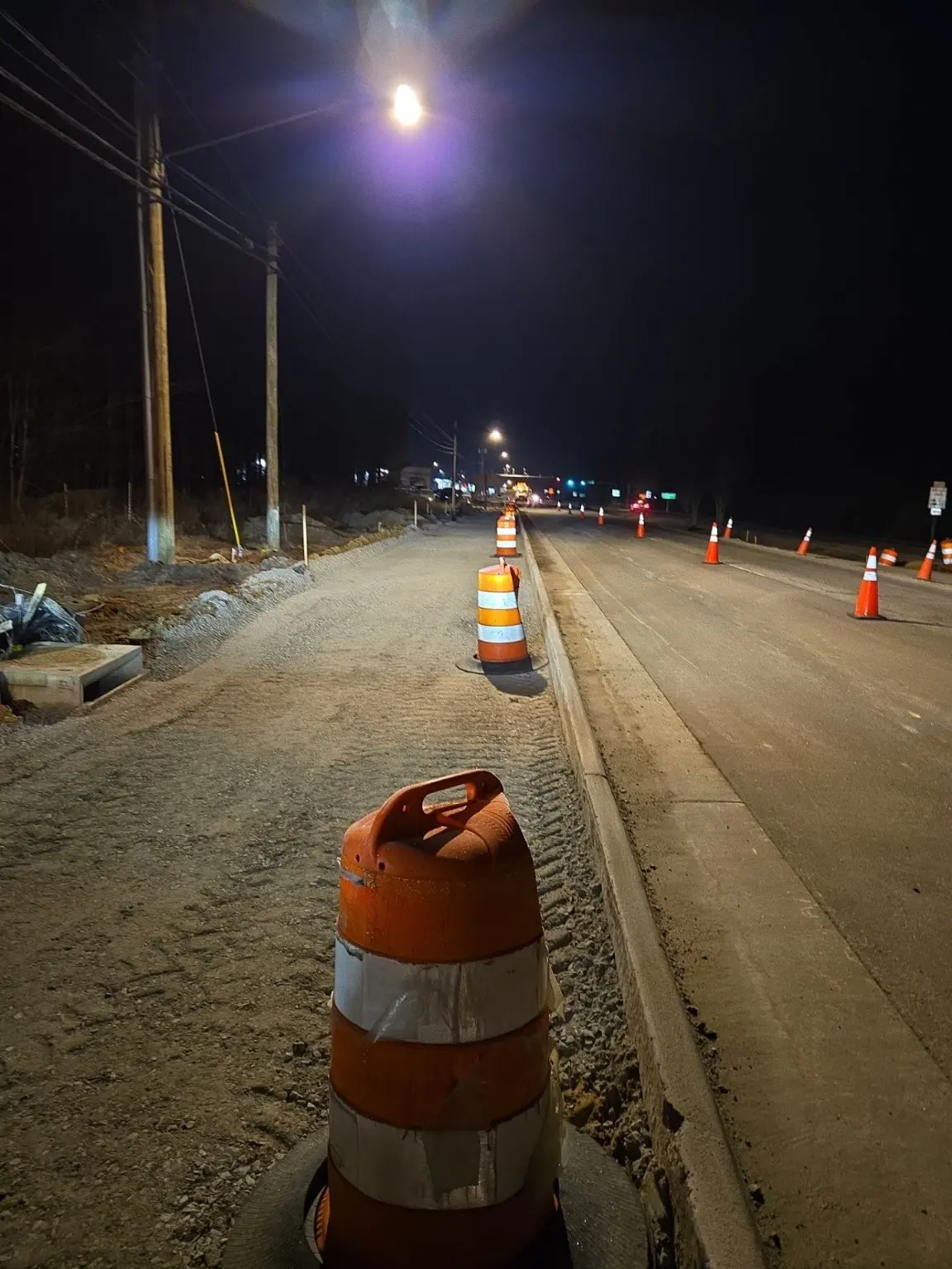 An aggregate roadside work area and concrete curb marked by traffic barrels at night.